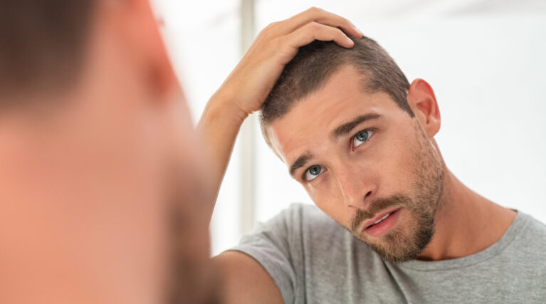 Young man checking hair in mirror Young unshaven man looking at mirror in bathroom at home. Handsome guy looking at his face in mirror, checking hair and hairline. Man in pijamas concerned with hair loss.