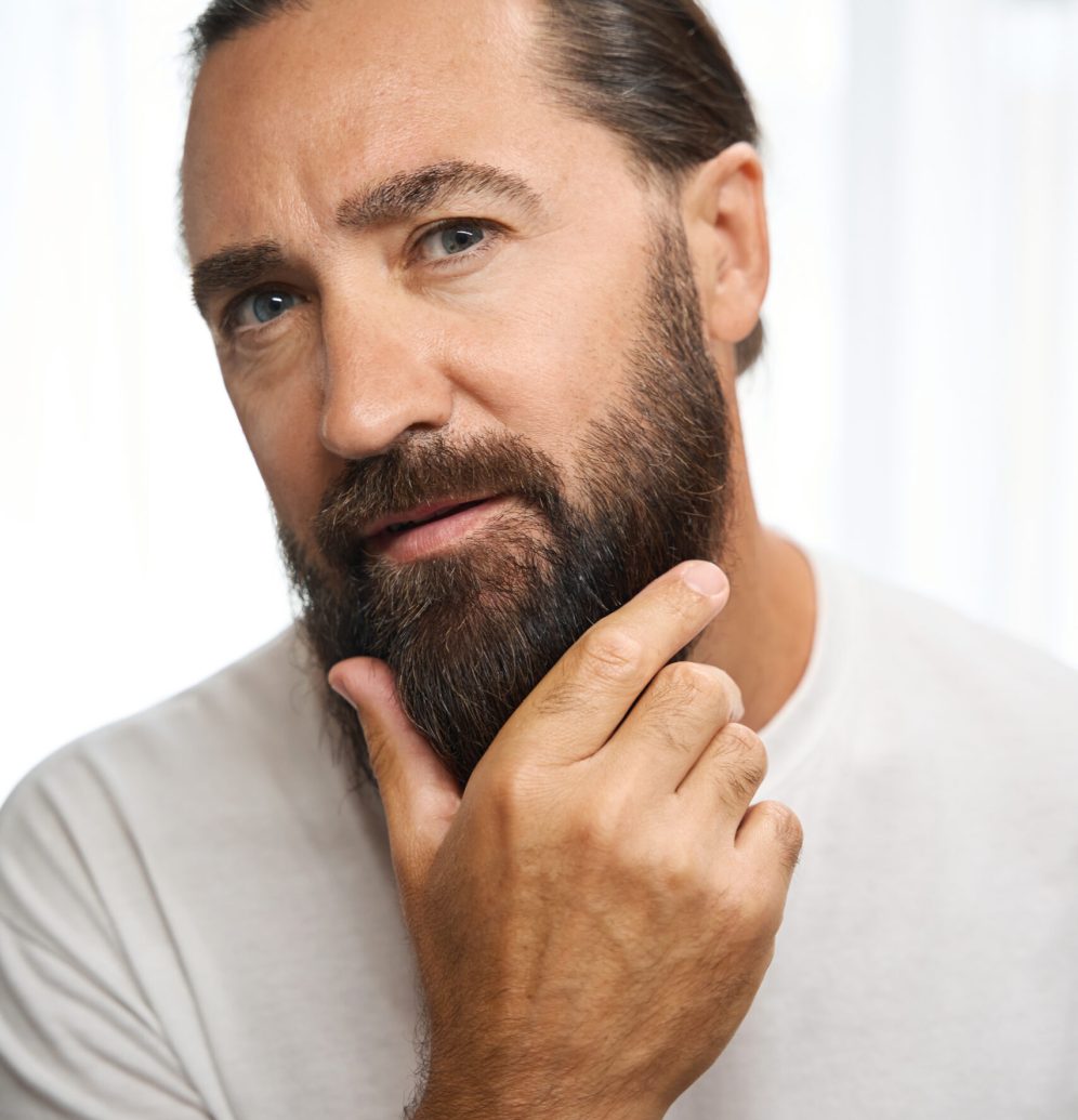 Cute male examines his thick beard, a man in white clothes